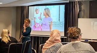 Facilitator engaging a group of early childhood educators during a Phoenix Support workshop, with a presentation slide in the background reading 'Say what you see and ask a question'.
